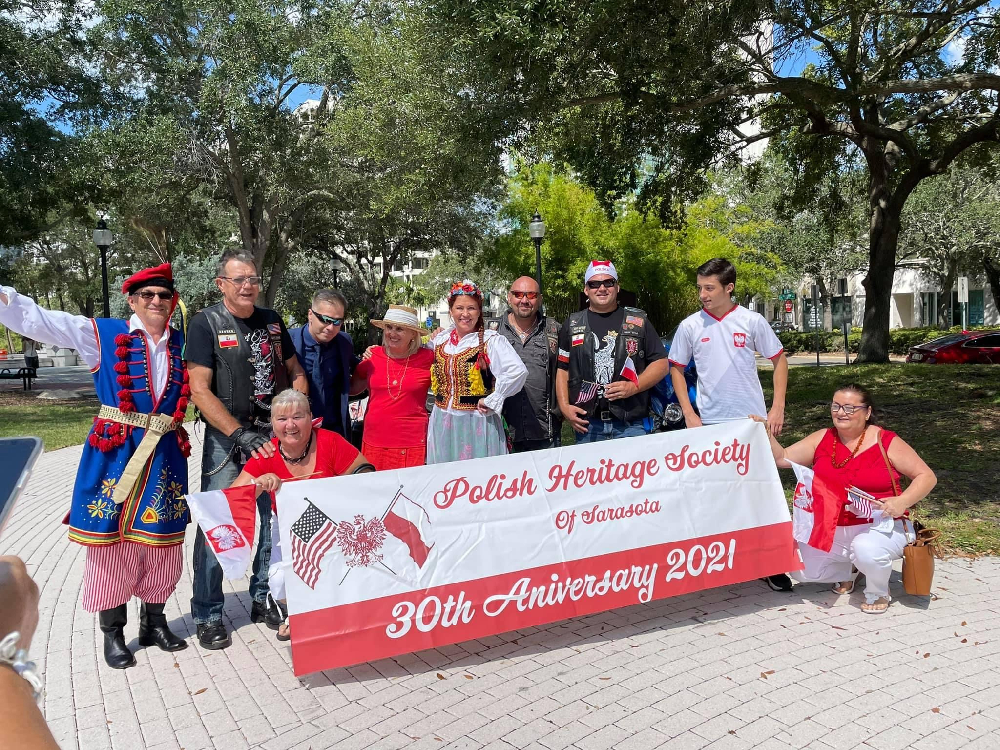 Uczestnicy spotkania Stowarzyszenia pozują na świeżym powietrzu, trzymając flagę Polski. Widzimy grupę ludzi o różnym wieku, ubranych w stroje reprezentujące ich polskie korzenie.